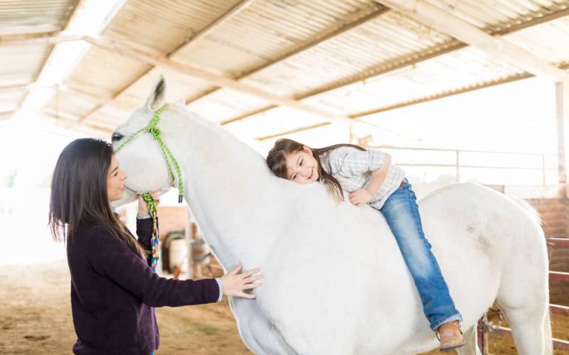 Bareback rider hugging her white horse while mother standing in stable